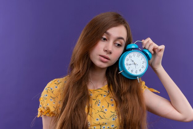 Задумчивая молодая девушка держит будильник на изолированной фиолетовой стене