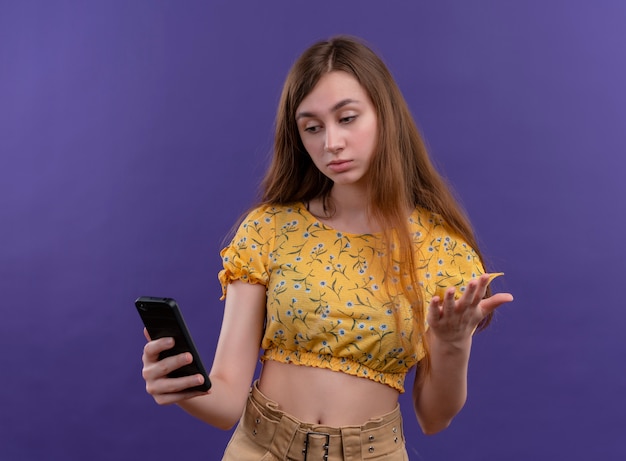 Задумчивая молодая девушка держит мобильный телефон и смотрит на него на изолированной фиолетовой стене