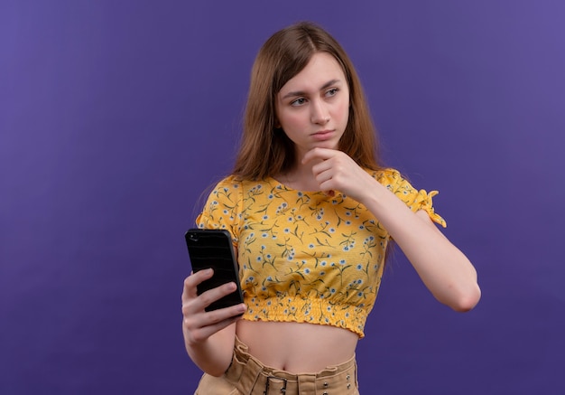 Задумчивая молодая девушка держит мобильный телефон и кладет руку на подбородок на изолированной фиолетовой стене с копией пространства