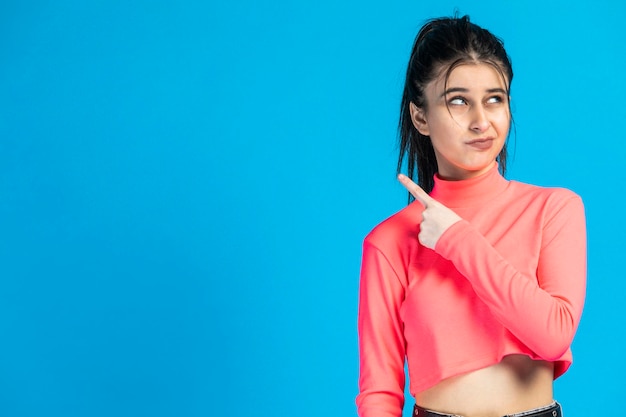 Thoughtful young girl wearing pink sweat and point finger to the side on blue background