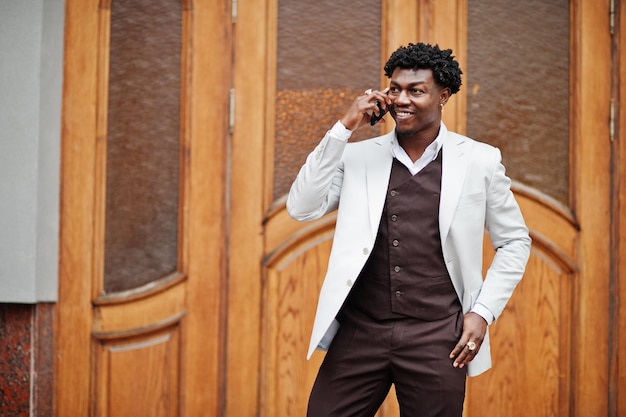 Вдумчивый молодой красивый афро-американский джентльмен в формальной одежде Черный стильный модельный мужчина в белой куртке разговаривает по телефону