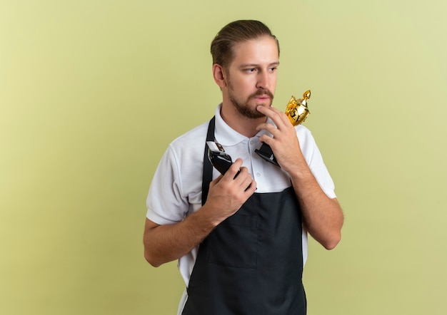 Thoughtful young handsome barber holding hair clippers and winner cup putting finger on chin looking at side isolated on olive green wall