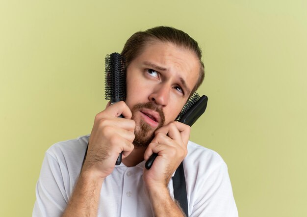 Thoughtful young handsome barber putting combs on face and looking up isolated on olive green wall