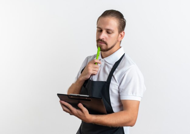 Thoughtful young handsome barber wearing uniform holding and looking at clipboard and touching his chin with pen isolated on white wall