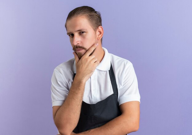 Thoughtful young handsome barber wearing uniform touching his chin and putting hand under elbow isolated on purple wall