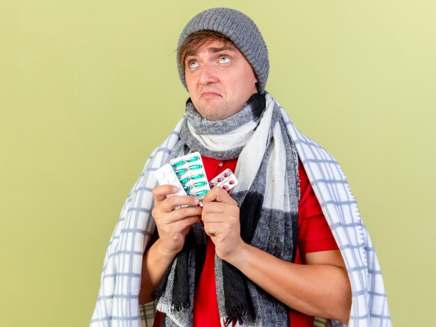 Thoughtful young handsome blonde ill man wearing winter hat and scarf wrapped in plaid holding packs of medical pills looking up isolated on olive green wall