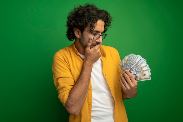 Thoughtful young handsome caucasian man wearing glasses holding and looking at money keeping hand on mouth isolated on green background with copy space