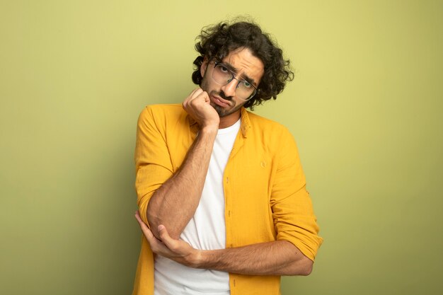 Thoughtful young handsome caucasian man wearing glasses looking at camera putting hand on chin isolated on olive green background with copy space
