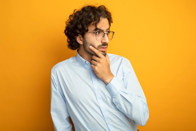 Thoughtful young handsome caucasian man wearing glasses looking at side holding chin isolated on orange background with copy space