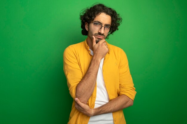 Free photo thoughtful young handsome caucasian man wearing glasses touching chin  isolated on green wall with copy space