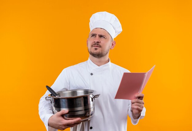 Вдумчивый молодой красивый повар в униформе шеф-повара держит котел и блокнот, глядя на изолированное оранжевое пространство