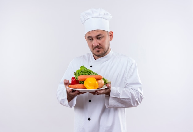 Thoughtful young handsome cook in chef uniform holding plate with vegetables looking at them on isolated white space