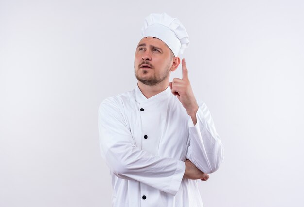 Thoughtful young handsome cook in chef uniform looking up with raised finger on isolated white space