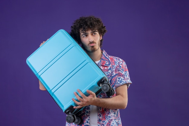Вдумчивый молодой красивый кудрявый путешественник мужчина держит чемодан, глядя на правую сторону на изолированной фиолетовой стене с копией пространства