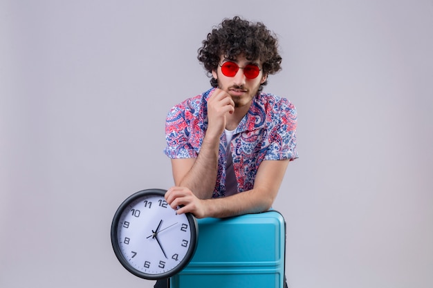 Thoughtful young handsome curly traveler man wearing sunglasses and holding clock and putting arms on suitcase with hand on chin on isolated white wall with copy space