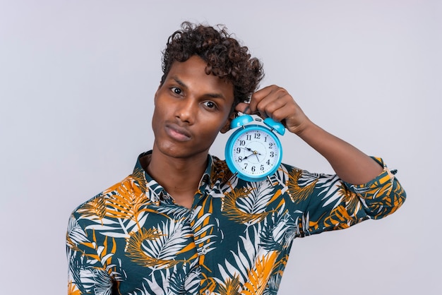 Thoughtful young handsome dark-skinned man with curly hair holding alarm clock listening to clock ticking sound on a white background