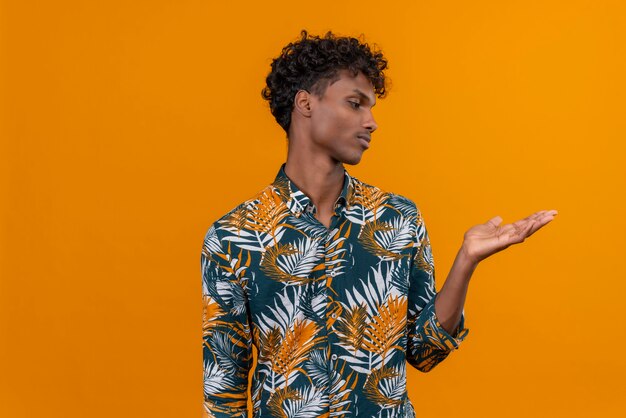 Задумчивый молодой красивый темнокожий мужчина с вьющимися волосами в рубашке с принтом листьев смотрит на свою ладонь на оранжевом фоне