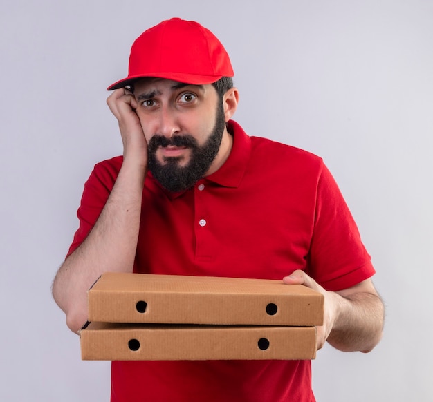 빨간 유니폼과 모자를 입고 사려 깊은 젊은 잘 생긴 배달 남자 똑바로보고 피자 상자를 들고 흰 벽에 고립 된 뺨에 손을 넣어
