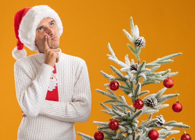 Thoughtful young handsome guy wearing christmas hat and santa claus tie standing near decorated christmas tree putting hand on chin looking up isolated on orange background