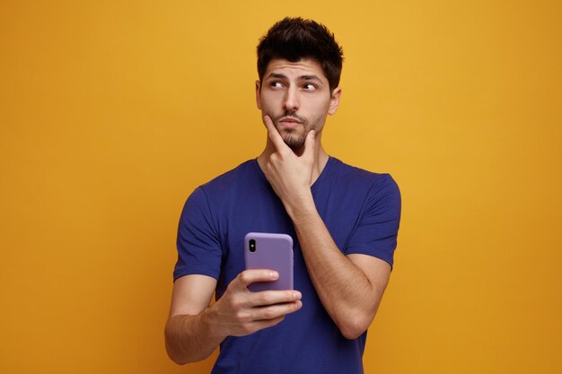 Thoughtful young handsome man holding mobile phone keeping hand on chin looking at side on yellow background