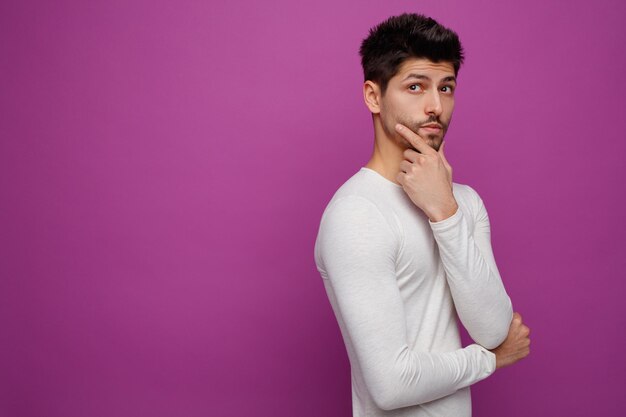 Thoughtful young handsome man standing in profile view putting hand on chin looking up on purple background with copy space