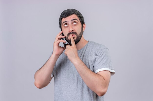 Thoughtful young handsome man talking on phone putting finger on lips and looking up isolated on white wall