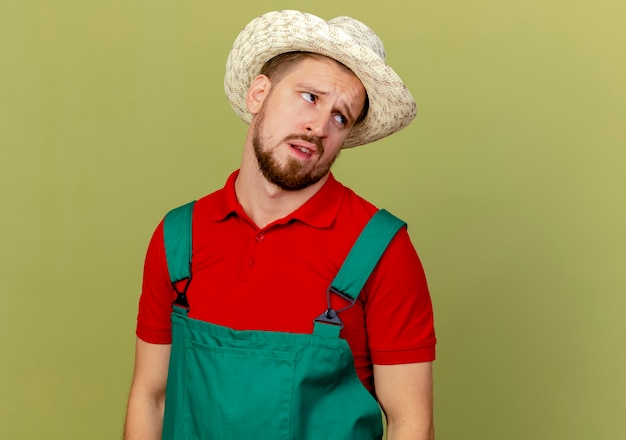 Giovane giardiniere slavo bello premuroso in uniforme e cappello che esaminano lato isolato sulla parete verde oliva con lo spazio della copia