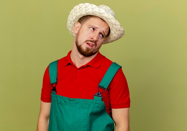 Задумчивый молодой красивый славянский садовник в униформе и шляпе смотрит в сторону, изолированную на оливково-зеленой стене с копией пространства