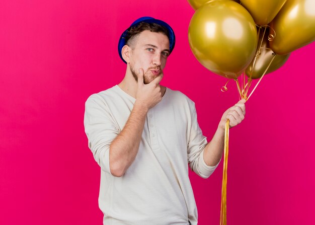 Thoughtful young handsome slavic party guy wearing party hat holding balloons looking at camera touching chin isolated on crimson background with copy space