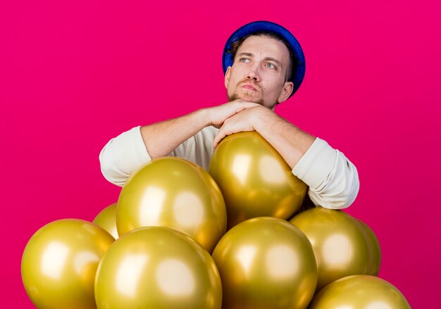 Thoughtful young handsome slavic party guy wearing party hat standing behind balloons putting hands on one of them looking at side isolated on crimson background