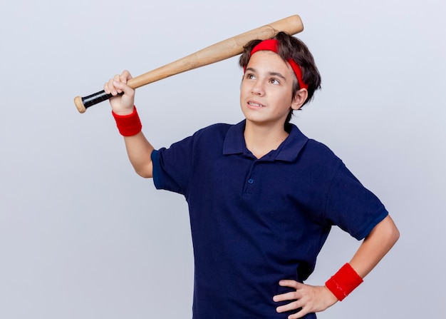 Вдумчивый молодой красивый спортивный мальчик с головной повязкой и браслетами с зубными скобами, держа руку на талии, глядя вверх, касаясь головы бейсбольной битой, изолированной на белой стене