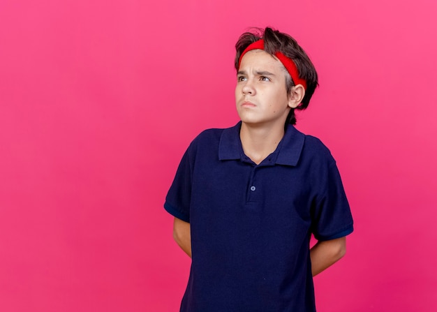 Thoughtful young handsome sporty boy wearing headband and wristbands with dental braces keeping hands behind back looking at side isolated on crimson background with copy space