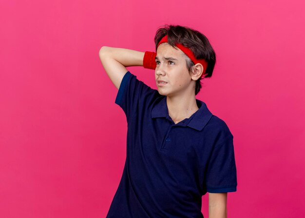 Thoughtful young handsome sporty boy wearing headband and wristbands with dental braces looking up keeping hand behind head isolated on pink wall with copy space