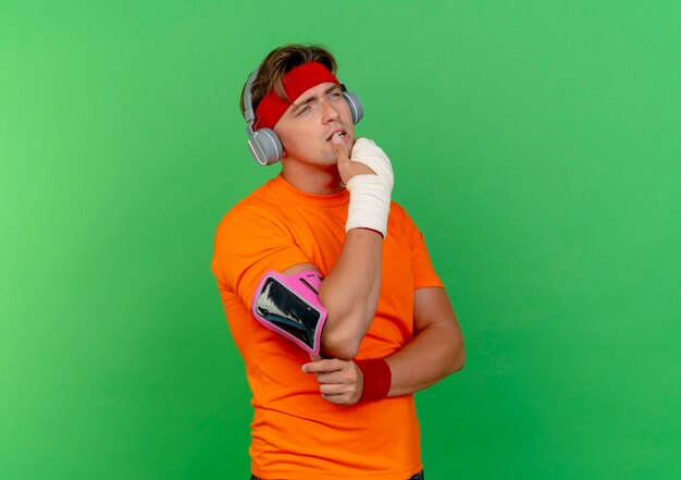 Thoughtful young handsome sporty man wearing headband and wristbands and headphones and phone armband with wrist wrapped with bandage putting finger on lip looking straight