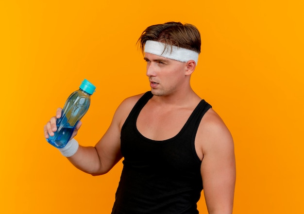 Free photo thoughtful young handsome sporty man wearing headband and wristbands holding water bottle looking straight isolated on orange wall