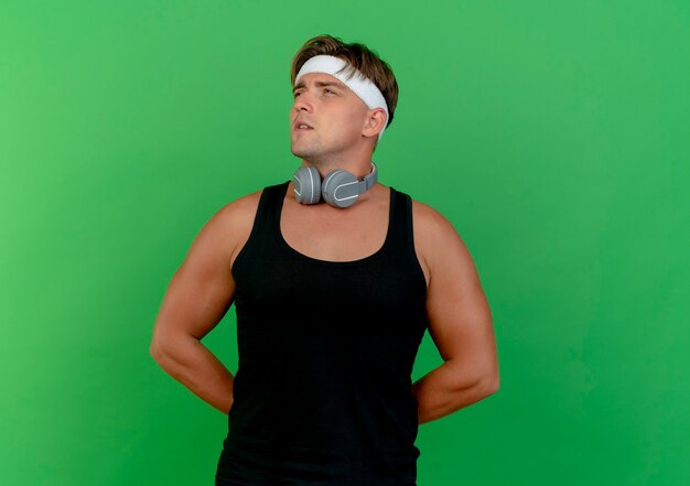 Thoughtful young handsome sporty man wearing headband and wristbands with headphones on neck putting hands behind back looking at side isolated on green wall