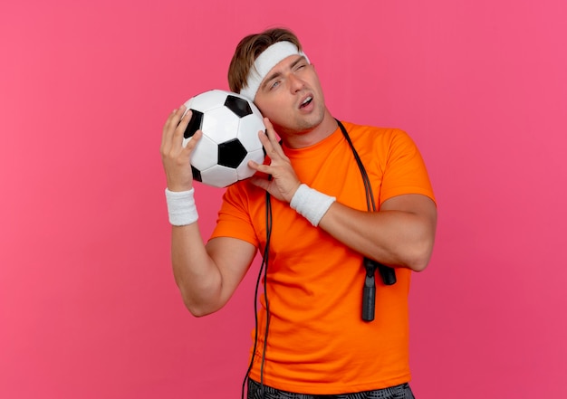 Вдумчивый молодой красивый спортивный мужчина с головной повязкой и браслетами со скакалкой на шее держит футбольный мяч, глядя в сторону, изолированную на розовой стене