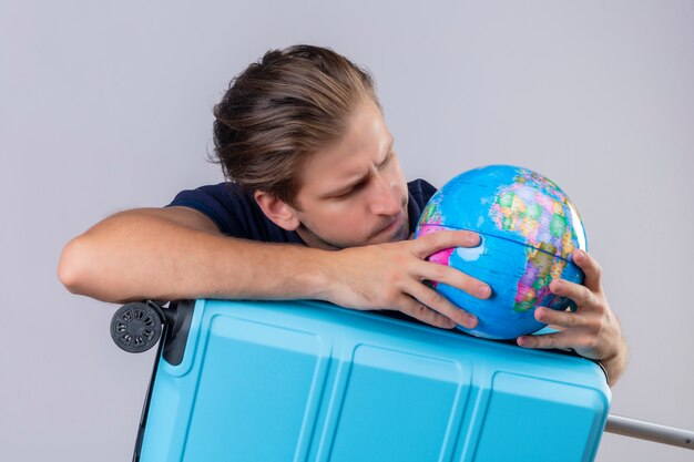 Thoughtful young handsome traveler guy standing with suitcase holding globe looking at it with serious face over white background