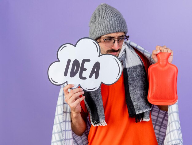 Thoughtful young ill man wearing glasses winter hat and scarf wrapped in plaid holding idea bubble and hot water bag looking at hot water bag isolated on purple wall with copy space