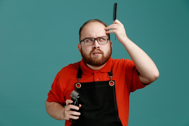 Free photo thoughtful young male barber wearing glasses red shirt and barber apron holding hair clipper and teaser comb looking at side while touching head isolated on blue background