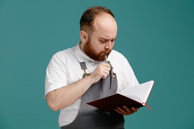 Giovane barbiere maschio premuroso che indossa una camicia bianca e grembiule da barbiere tenendo e guardando il blocco note mentre si tocca il labbro con la penna isolato su sfondo blu