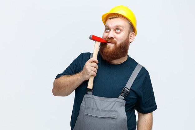 Thoughtful young male construction worker wearing safety helmet and uniform touching lip with hammer looking up isolated on white background