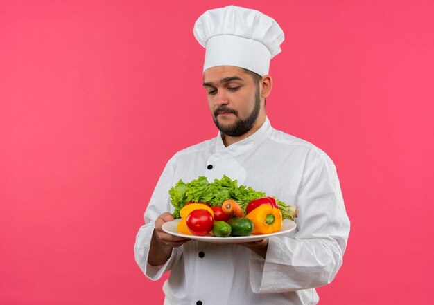 Thoughtful young male cook in chef uniform holding and looking at plate of vegetables isolated on pink space 