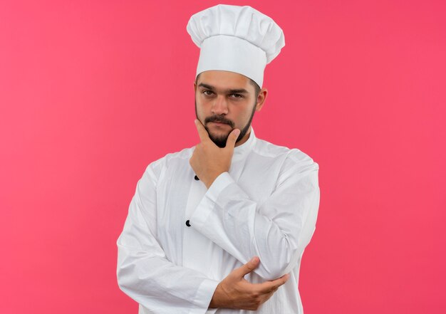 Вдумчивый молодой мужчина-повар в униформе шеф-повара положил руку на подбородок и локоть, изолированные на розовом пространстве