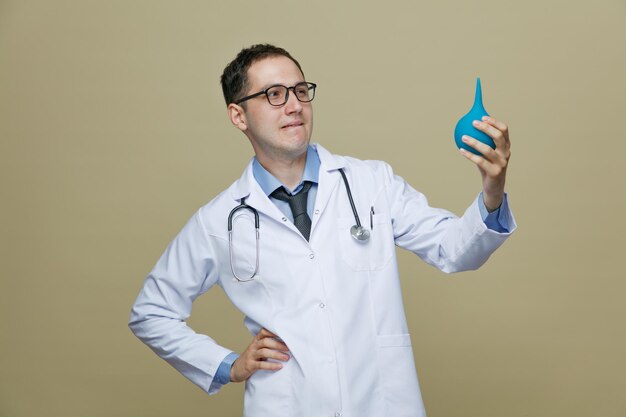 Thoughtful young male doctor wearing glasses medical robe and stethoscope around neck holding enema looking at it while keeping hand on waist and biting lip isolated on purple background