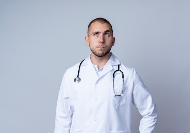 Thoughtful young male doctor wearing medical robe and stethoscope around his neck looking up isolated on white wall