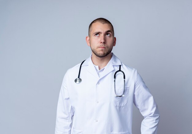Вдумчивый молодой мужчина-врач в медицинском халате и стетоскопе на шее, глядя вверх изолирован на белой стене
