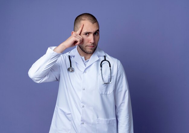 Foto gratuita giovane medico maschio premuroso che indossa veste medica e stetoscopio intorno al collo che mette il dito sulla testa isolata sulla parete viola