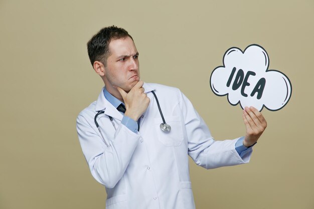 thoughtful young male doctor wearing medical robe and stethoscope around neck holding idea bubble looking at it while keeping hand on chin isolated on olive green background