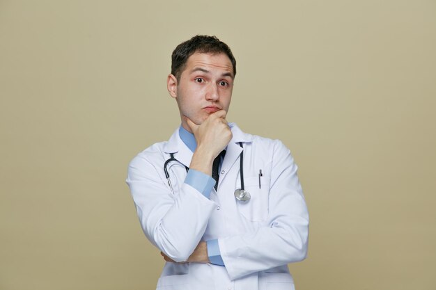 Thoughtful young male doctor wearing medical robe and stethoscope around neck looking at side while keeping hand on chin isolated on olive green background